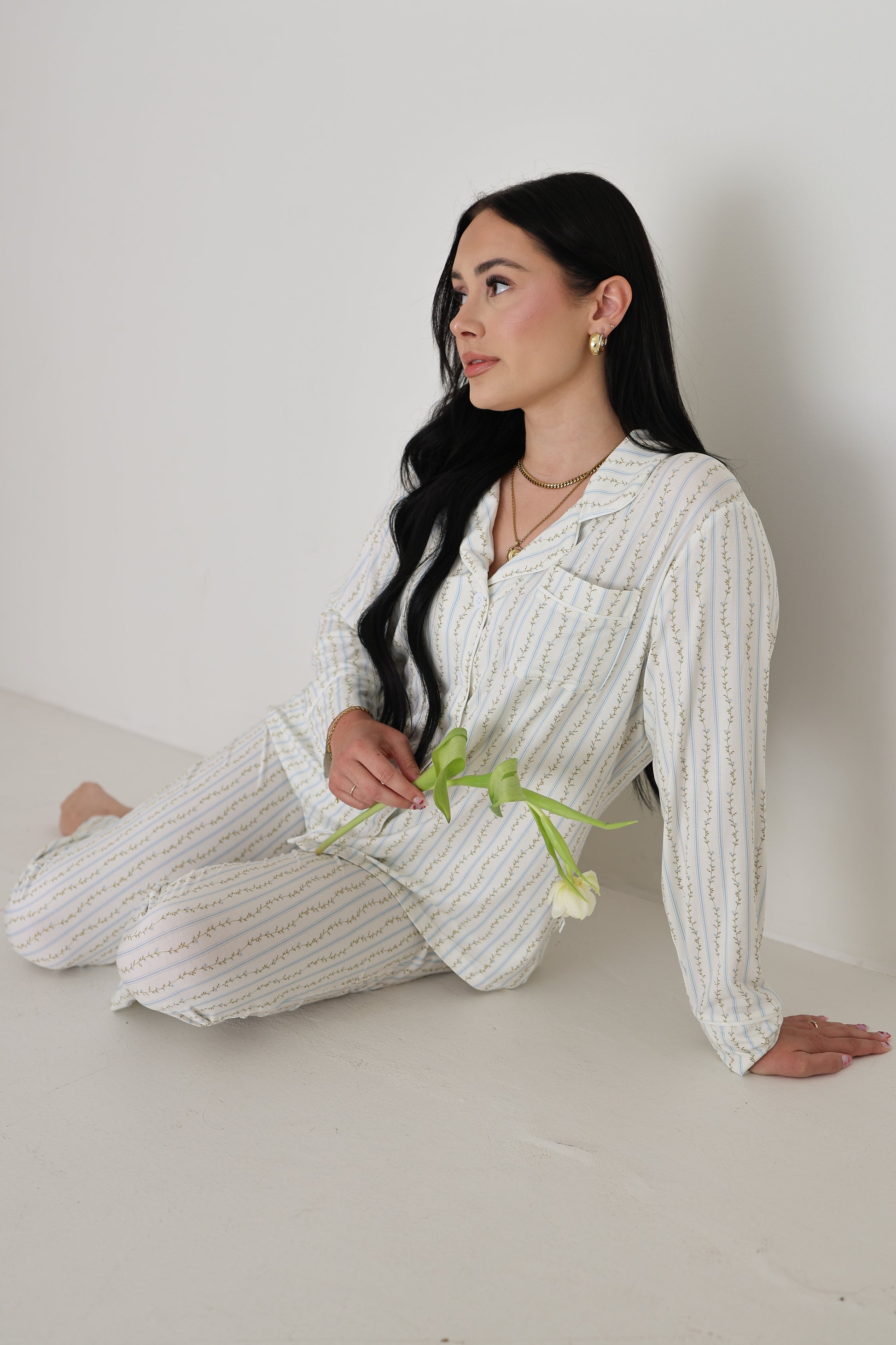A woman with long dark hair sits on the floor in a forever french baby Women's Bamboo Button Up Lounge Set | Dainty Vines, holding a white tulip and enjoying the comfort of this hypo-allergenic bamboo loungewear.