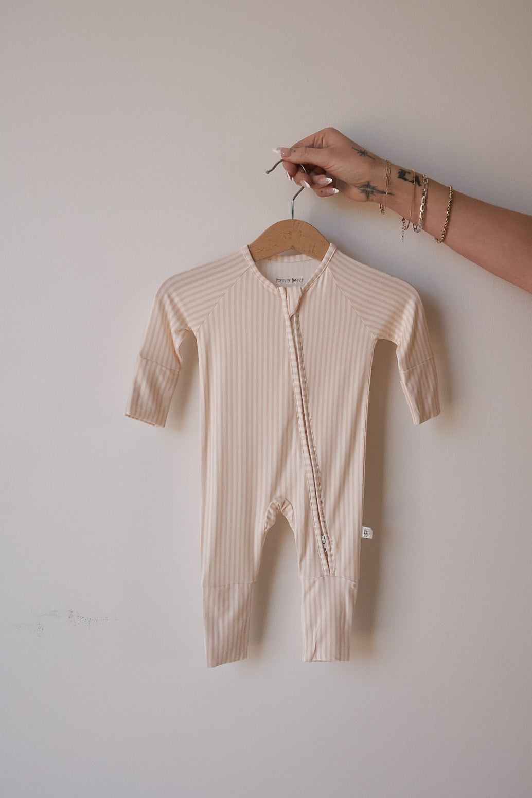A hand holds the forever french baby Bamboo Zip Pajamas | Sadie Stripe—beige and white striped, long-sleeve baby sleepwear with a breathable fabric and diagonal neck-to-leg zipper—against a plain light-colored wall.