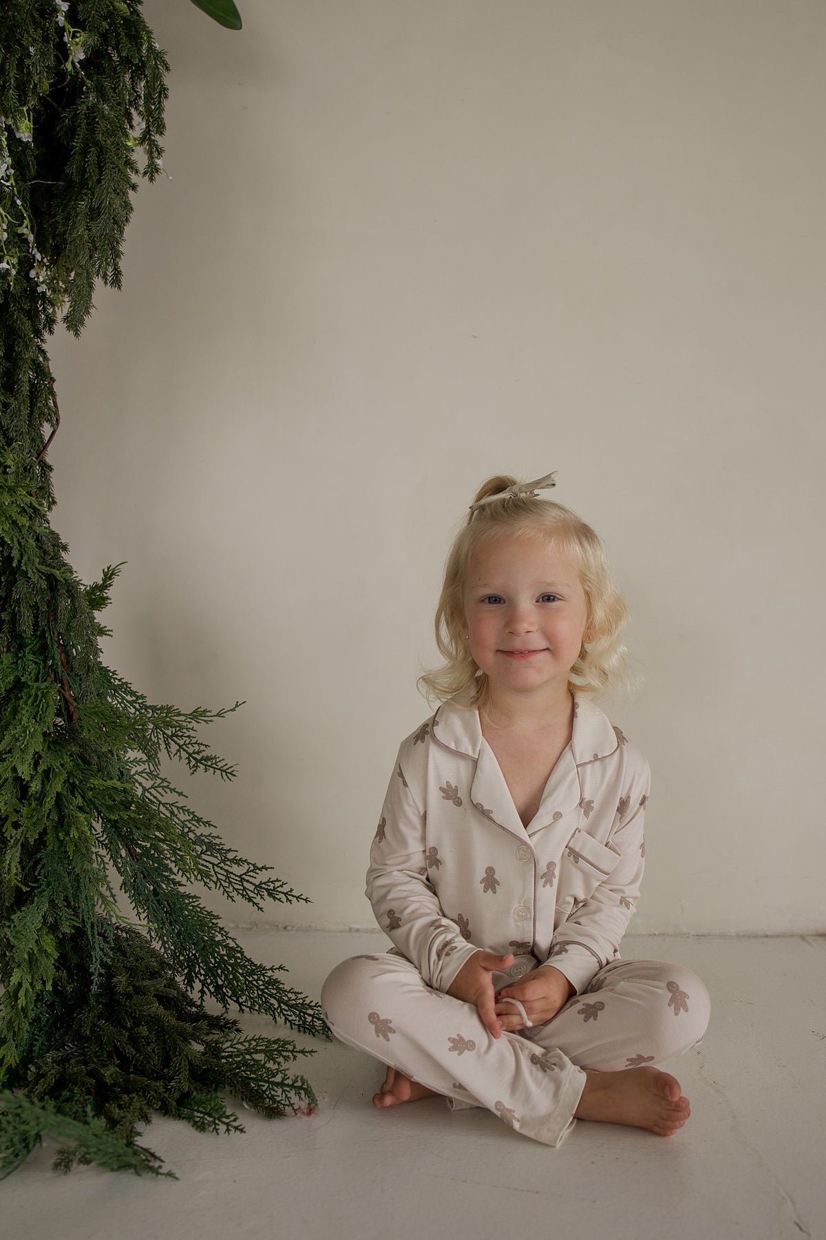 A young child sits cross-legged next to a pine bough wearing a Girl’s Bamboo Button Up Lounge Set | Gingersnap.