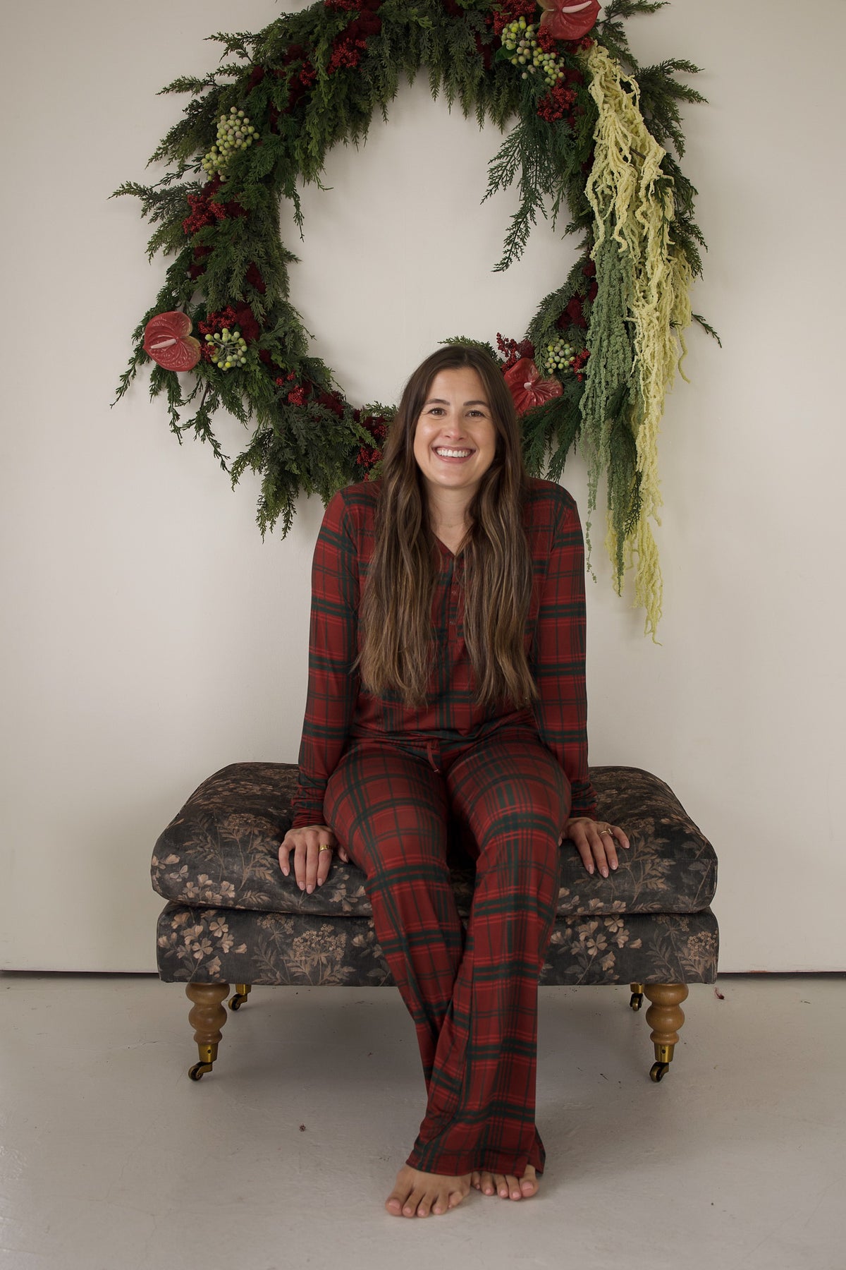 A smiling woman sits on a patterned ottoman wearing Women’s Long Sleeve Bamboo Pajama | Christmas Plaid in front of a large holiday wreath.