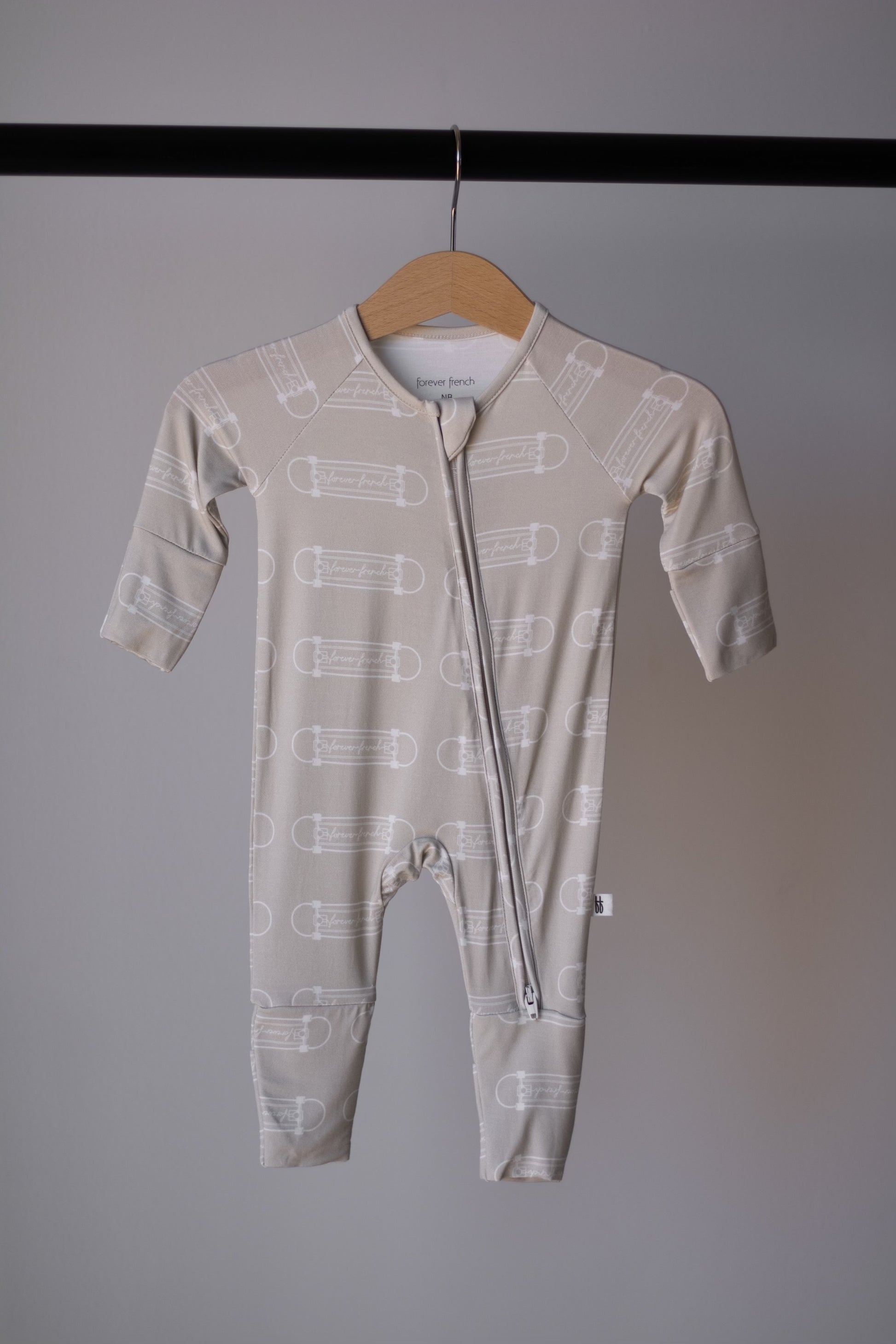 The forever french baby Bamboo Zip Pajamas | Sk8er Boy is a beige infant onesie with a white safety pin pattern, long sleeves and legs, made from breathable sleepwear fabric and featuring a diagonal zipper closure for easy changes.