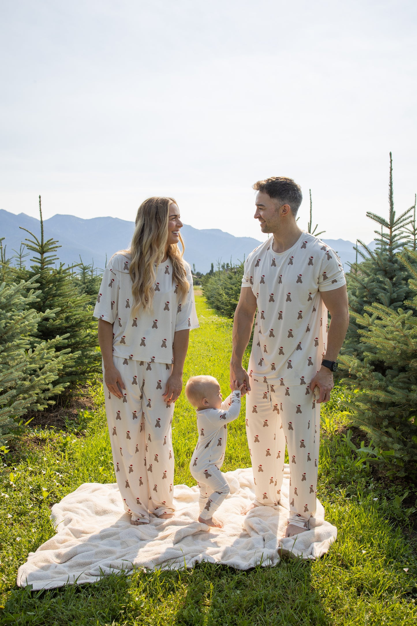 A man wearing forever french baby's Men's Short Sleeve Pajamas in the Santa Teddy print stands on a blanket among rows of evergreen trees with a woman and toddler, all dressed in matching hypoallergenic sleepwear. The adults smile at each other while holding the child's hands, surrounded by a bright outdoor setting with mountains in the background—perfectly capturing a cozy family moment in comfortable bamboo pajamas.
