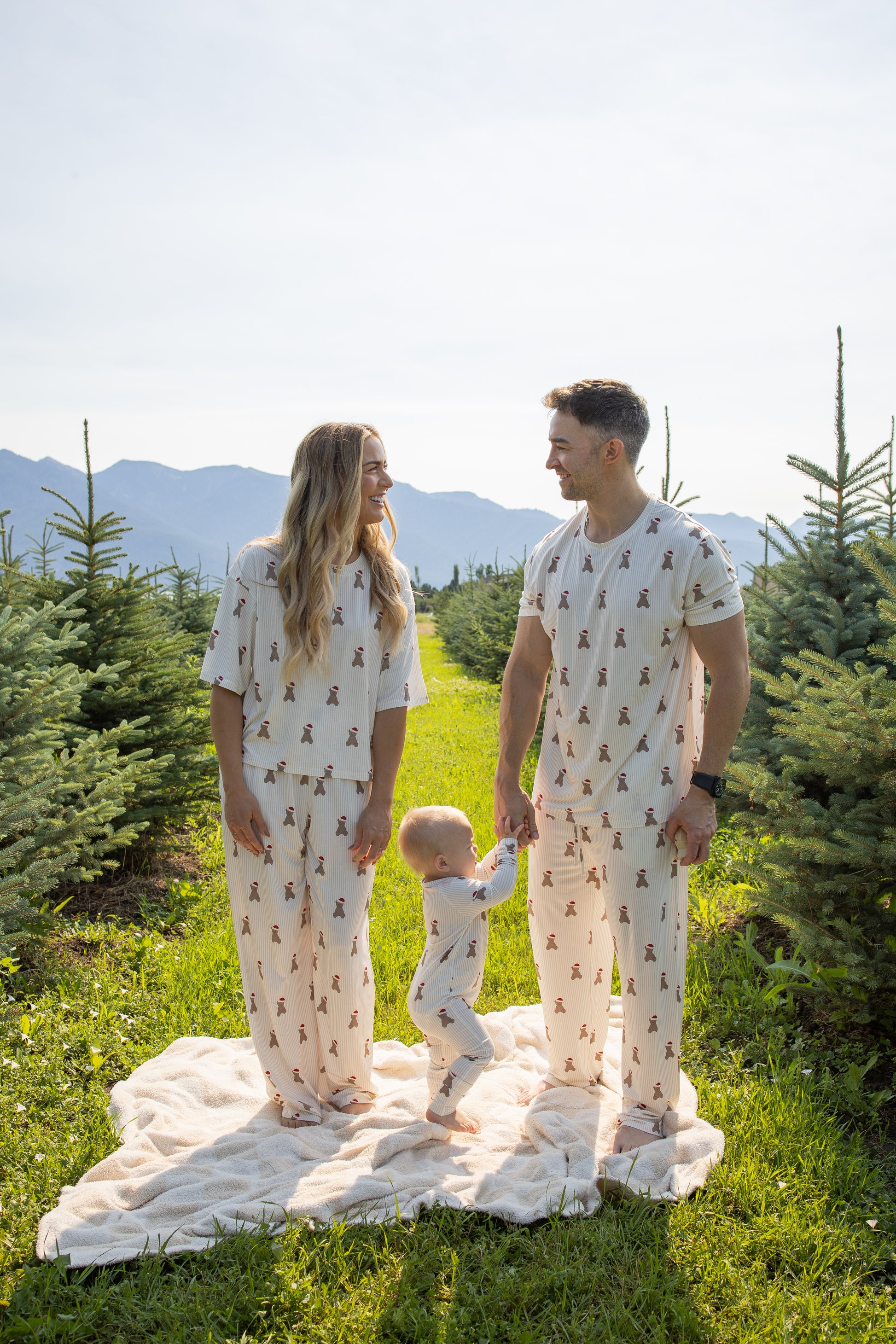 A man wearing forever french baby's Men's Short Sleeve Pajamas in the Santa Teddy print stands on a blanket among rows of evergreen trees with a woman and toddler, all dressed in matching hypoallergenic sleepwear. The adults smile at each other while holding the child's hands, surrounded by a bright outdoor setting with mountains in the background—perfectly capturing a cozy family moment in comfortable bamboo pajamas.