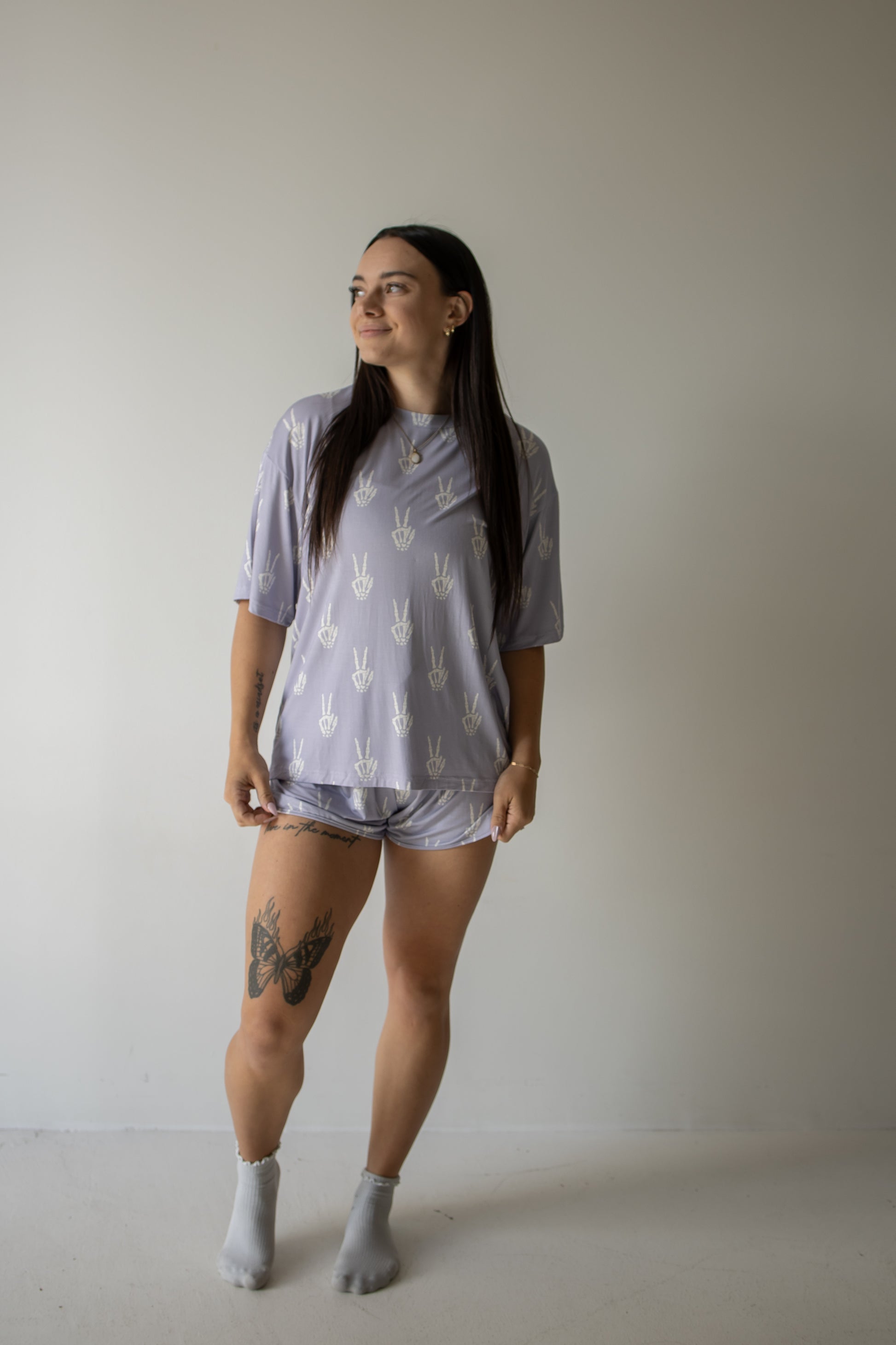 A woman with long dark hair stands against a plain light-colored wall, looking to her left. She is wearing the forever french baby Women’s Bamboo Short Set | Enchanted Skelly, a hypo-allergenic lavender pajama set with white patterns. She has a large butterfly tattoo on her thigh and is also wearing light gray socks.