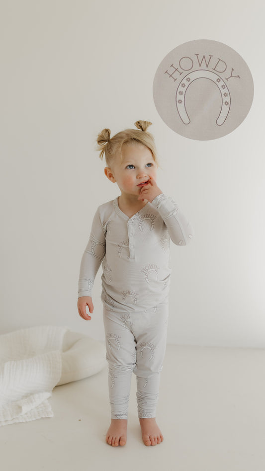 A toddler with pigtails stands barefoot on a light floor, wearing forever french baby Bamboo Two Piece Pajamas featuring horse patterns. She looks up, hand near her mouth, while a "Howdy" design with a horseshoe perfectly complements the playful theme.