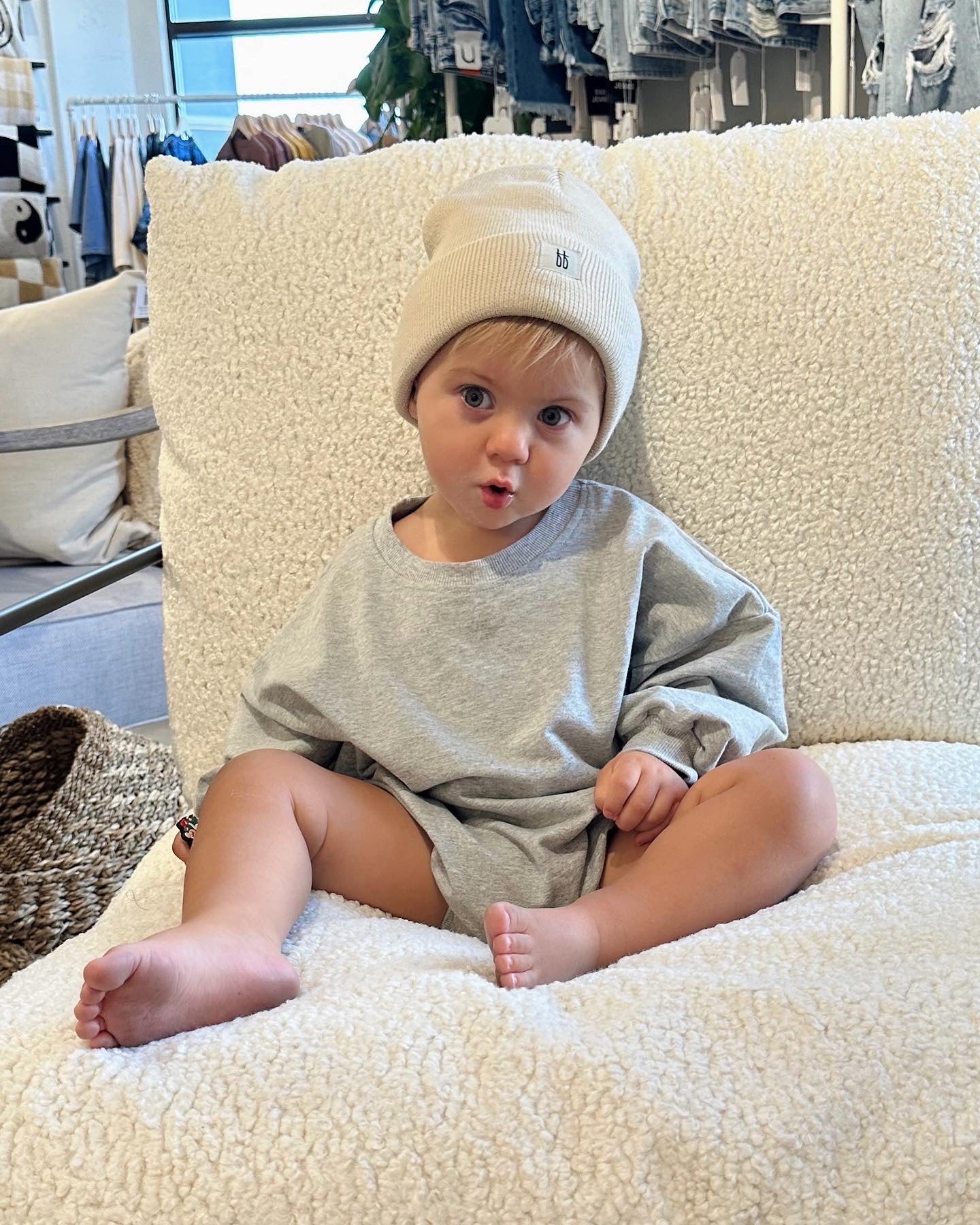 A toddler with short hair wearing a beige beanie and the oversized gray "Sweatshirt Romper | Stormi" from forever french baby, made from the softest material, sits on a fluffy chair with legs crossed. The child looks into the camera with a playful, surprised expression. The background shows a cozy, trendy room filled with various items.