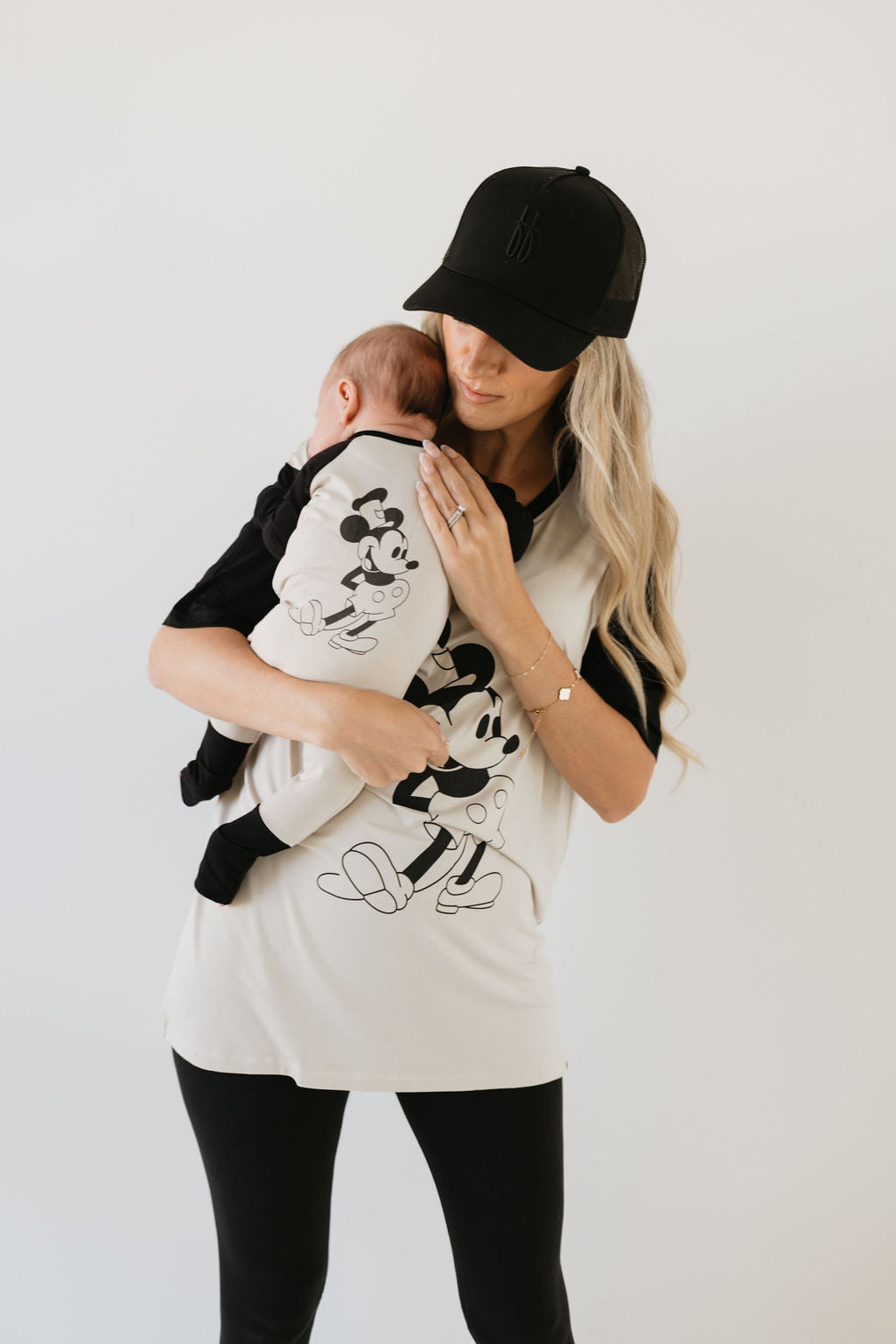 A woman with long blonde hair, wearing a black cap and leggings, holds a baby dressed in forever french baby's Bamboo Zip Pajamas in the Steamboat Willie design. She is also wearing a Mickey Mouse shirt and stands against a plain background.