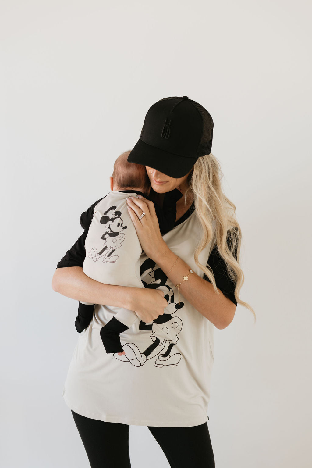 A person with long blonde hair and a black cap holds a baby, both dressed in forever french baby Bamboo Zip Pajamas showcasing Steamboat Willie designs. The pair smiles at each other against a plain background, radiating joy in their coordinated outfits.