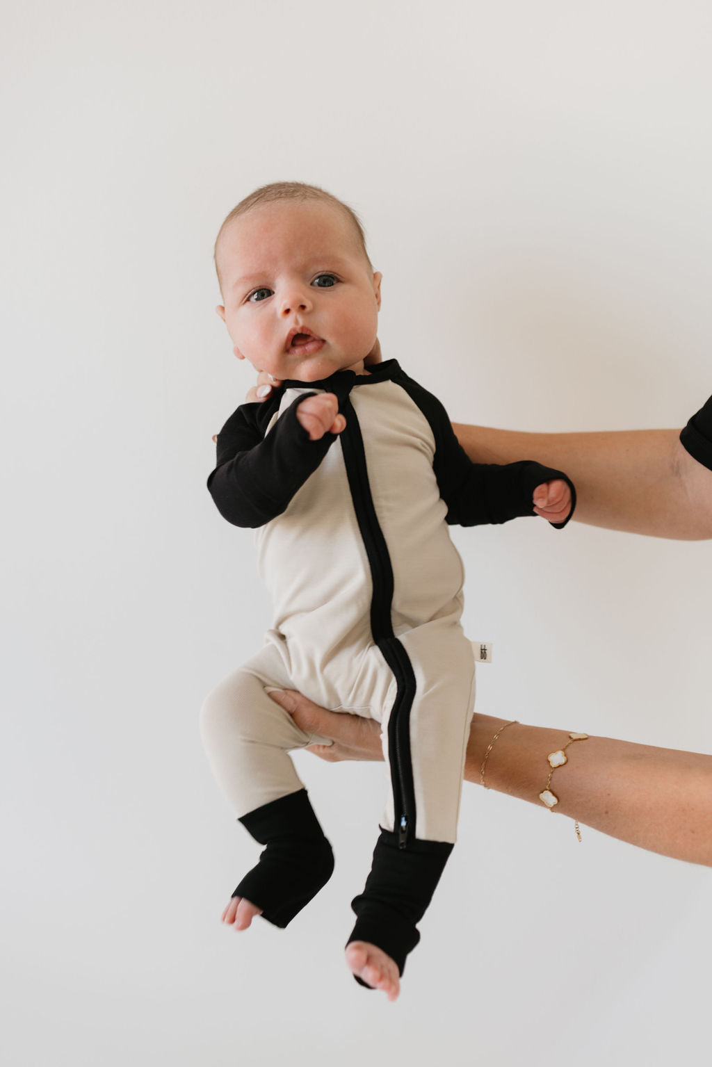 A baby wearing "Bamboo Zip Pajamas | Steamboat Willie" by forever french baby is held up against a white background. The beige and black onesie complements the child's calm and curious eyes, while a gentle hand provides support, ensuring comfort and care.