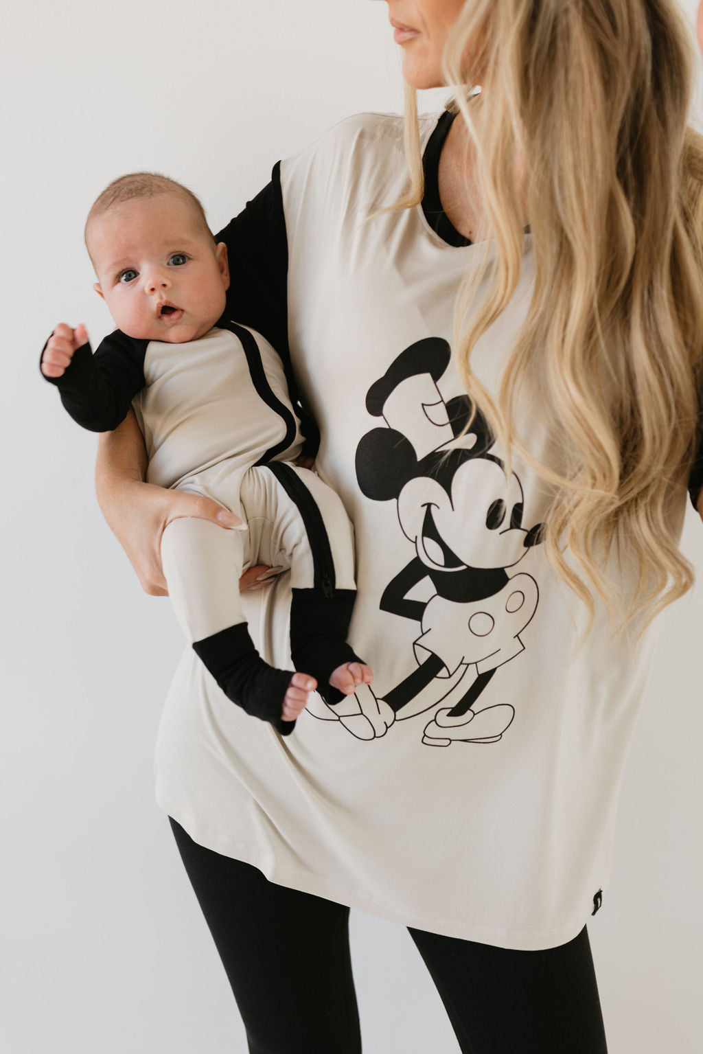 A woman with long blond hair holds a baby, both dressed in matching black and white outfits featuring a cartoon character. Her tunic boasts a large mouse illustration, while the baby is wearing "Bamboo Zip Pajamas | Steamboat Willie" from forever french baby, a breathable onesie perfect for sensitive skin.