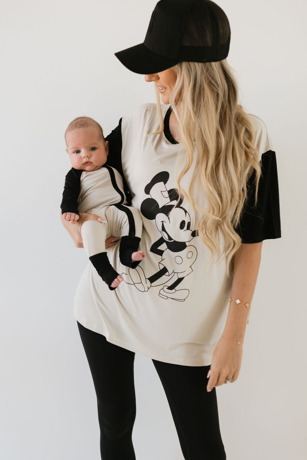 A woman with long, blonde hair in a baseball cap is holding a baby, both dressed in matching Bamboo Zip Pajamas featuring the Steamboat Willie design by forever french baby. The breathable material ensures comfort while the hypo-allergenic clothing is gentle against sensitive skin. The background is plain white.