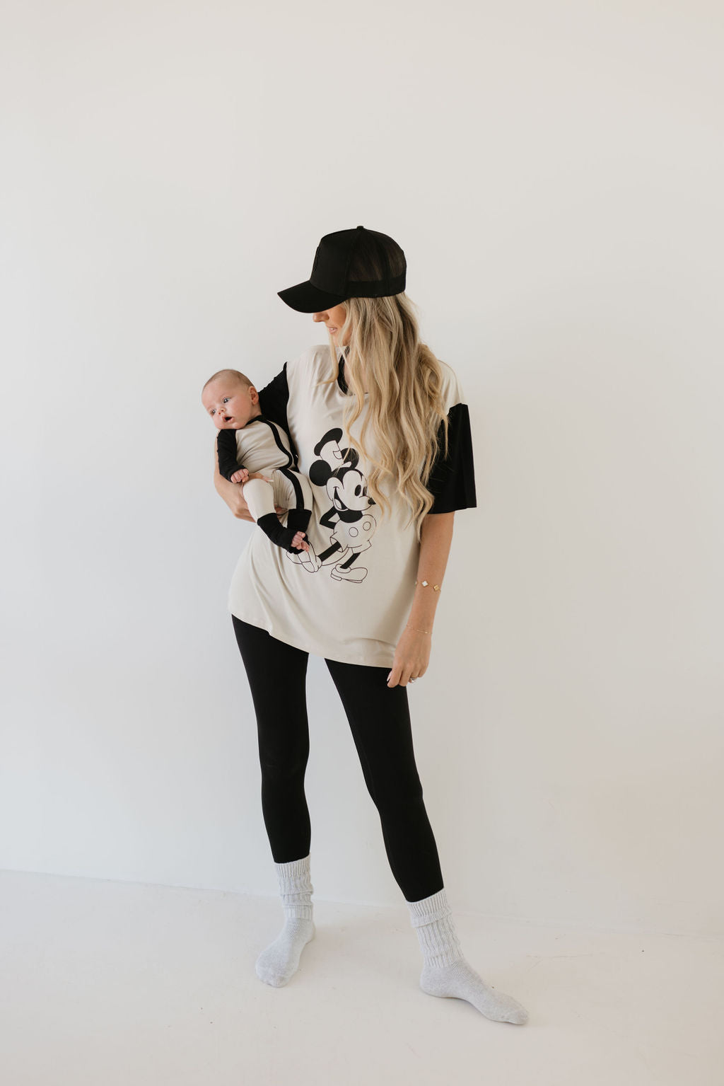 A woman with long blonde hair wearing a black cap, an oversized Mickey Mouse t-shirt, black leggings, and white socks cradles a baby dressed in breathable, hypo-allergenic **forever french baby** Bamboo Zip Pajamas in the Steamboat Willie design. The background is a plain white wall.