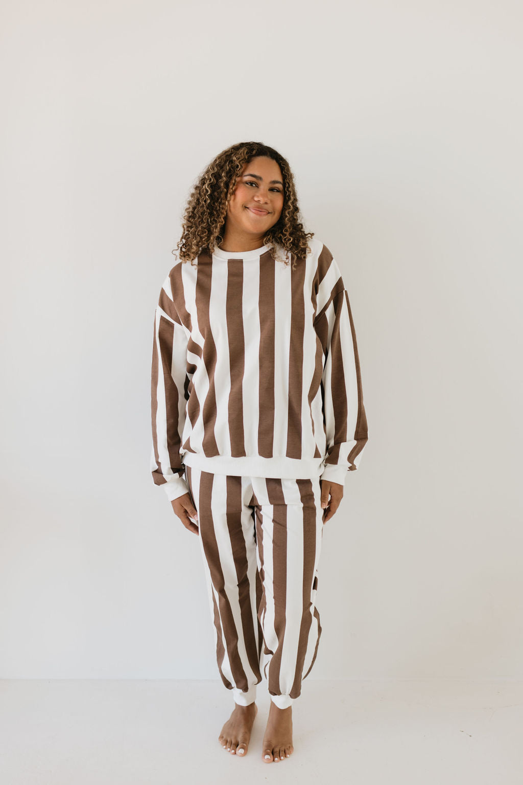 A person with curly hair is smiling, wearing the Adult Sweat Set from forever french baby called Coffee Shoppe, featuring matching brown and white striped sweater and pants. They stand barefoot against a plain white background.