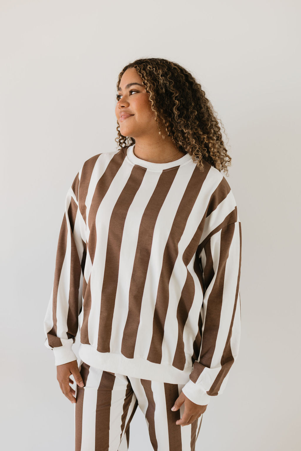 A person with curly hair wearing the forever french baby Adult Sweat Set in a chic brown and white vertical stripe glances to the side against a plain light background, radiating the relaxed vibe of a cozy coffee shoppe.