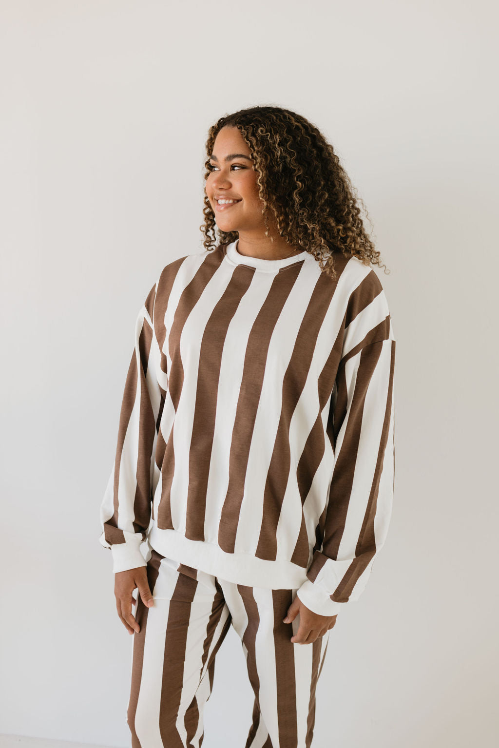 A person with curly hair smiles while wearing the stylish "Adult Sweat Set | Coffee Shoppe" by forever french baby, standing against a plain white background.