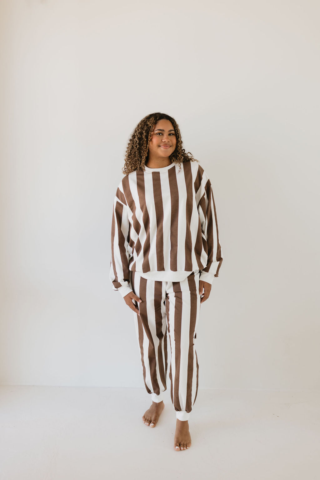 Against a plain white backdrop, a person with curly hair stands barefoot, donning the Adult Sweat Set | Coffee Shoppe by forever french baby. This matching ensemble showcases bold brown and white vertical stripes and includes both a long-sleeved shirt and pants.