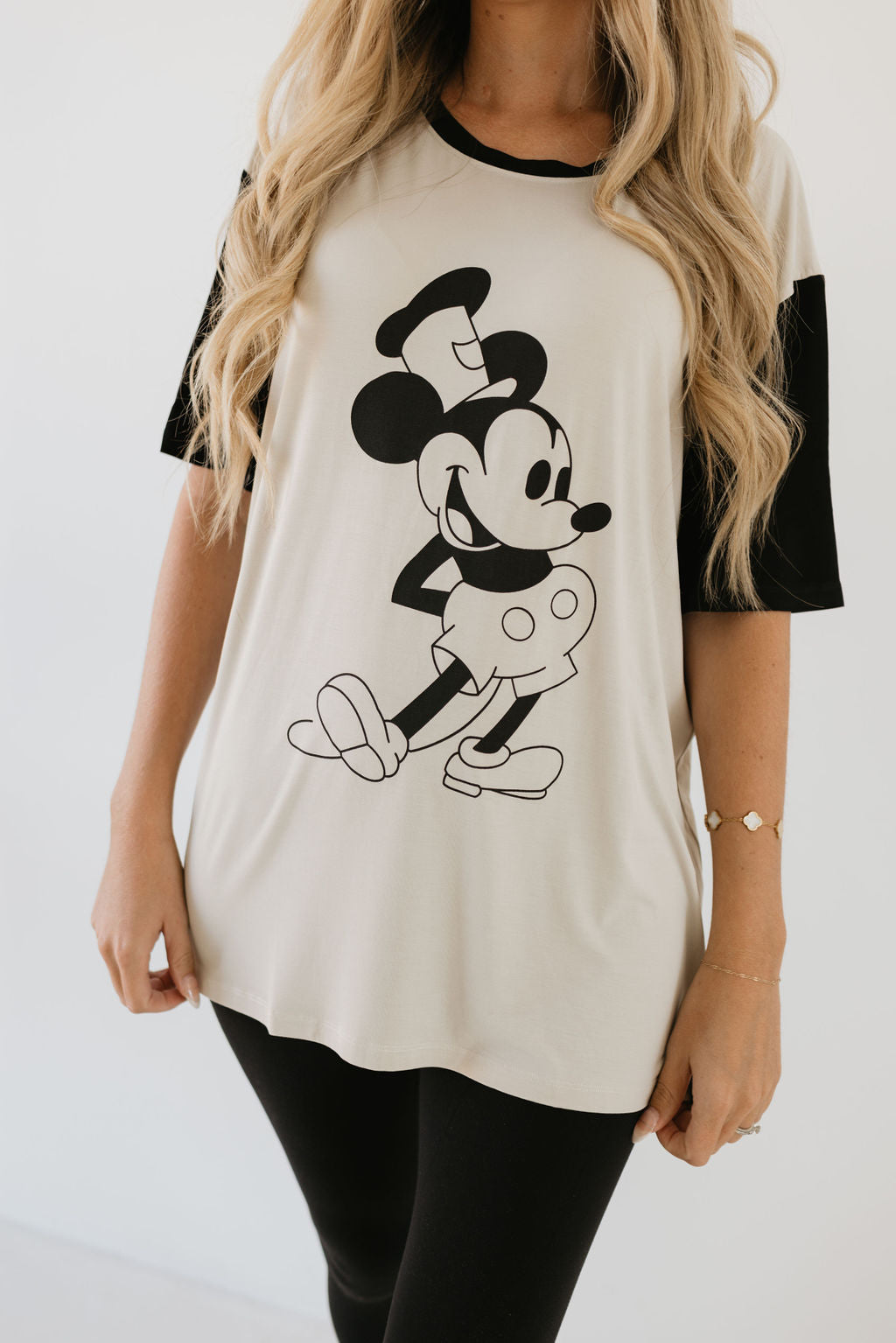 Someone with long, wavy hair is wearing a forever french baby Adult Bamboo Tee featuring a classic Steamboat Willie design, paired with black leggings. The character with large ears and gloves is depicted in a playful pose that captures the whimsy of early animation.