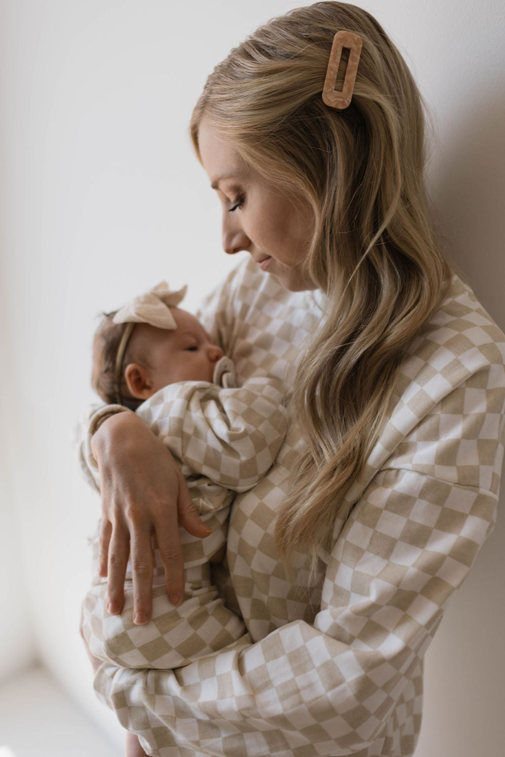 A woman with long blonde hair, wearing a hair clip and the Adult Long Sleeve Short Set in Chai Checkerboard by forever french baby, lovingly cradles a sleeping baby dressed in matching attire. The baby has a small bow headband, and both appear serene and content. The background is softly lit and minimalistic, capturing a perfect mama-and-me moment.