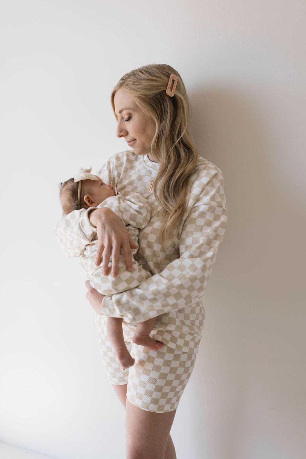 A woman with long blonde hair, clipped to the side, holds a baby close to her chest. Both are wearing coordinating sets from forever french baby in matching beige and white checkered outfits, with the woman dressed in the Adult Long Sleeve Short Set | Chai Checkerboard. The woman is looking down lovingly at the baby, who has a bow on their head. They are standing against a plain, light background.