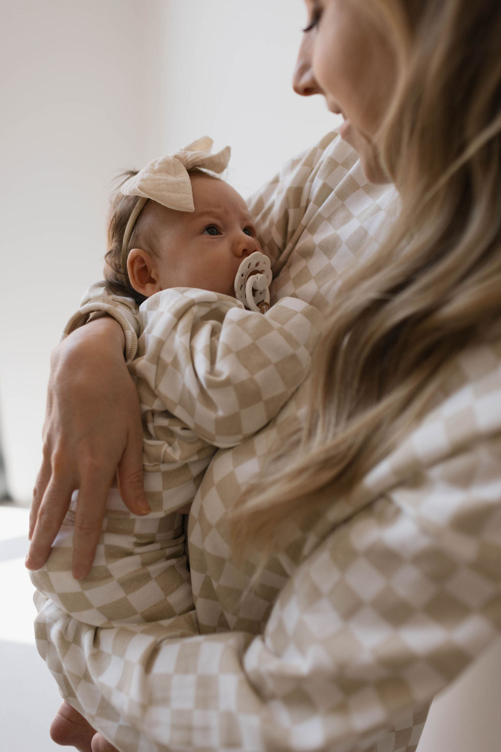 A woman cradles a baby dressed in a light checkered onesie with a pacifier and bow headband. Both are wearing matching Adult Long Sleeve Short Sets in the Chai Checkerboard design by forever french baby, sharing a tender mama-and-me moment as the woman looks down lovingly at the baby in a softly lit room.