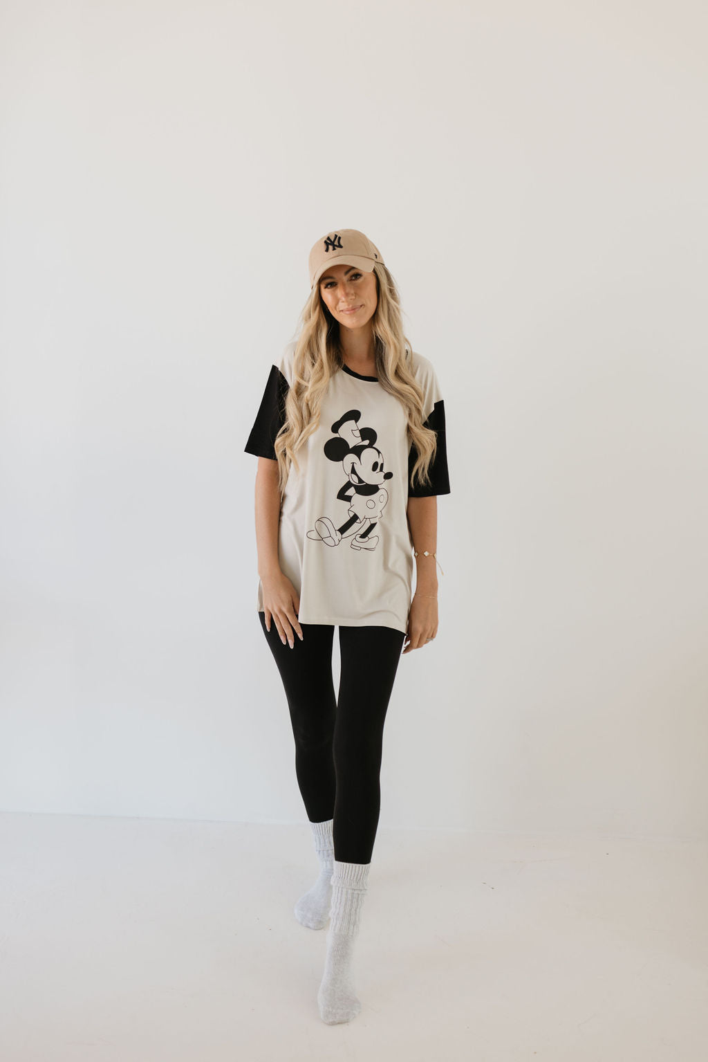 A person wearing a tan baseball cap, a white "Adult Bamboo Tee | Steamboat Willie" by forever french baby, black leggings, and gray socks stands against a plain white background.