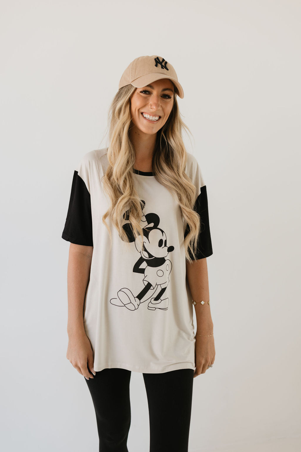 A woman with long blonde hair is smiling, dressed in a beige cap and a black and white "Adult Bamboo Tee | Steamboat Willie" by forever french baby, paired with black pants. She stands against a plain background, radiating charm in her comfortable attire.