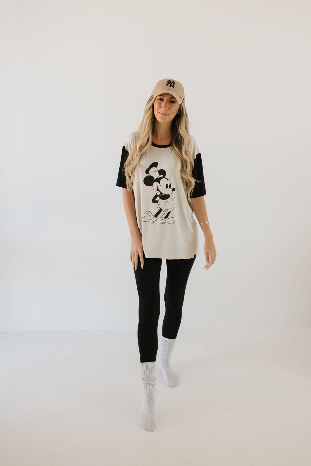 A woman with long hair stands against a plain background, wearing the beige Adult Bamboo Tee featuring Steamboat Willie from forever french baby. She pairs it with black leggings and gray socks, creating a casual yet stylish look.