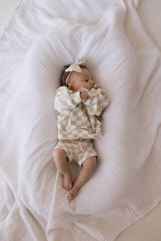 A baby lies on a crinkled white blanket, wearing the Children's Long Sleeve Short Set in Chai Checkerboard from forever french baby that matches mama-and-me styles. The baby has a bow headband and looks to the side while clutching part of the blanket, with the serene setting emphasizing their peaceful expression.