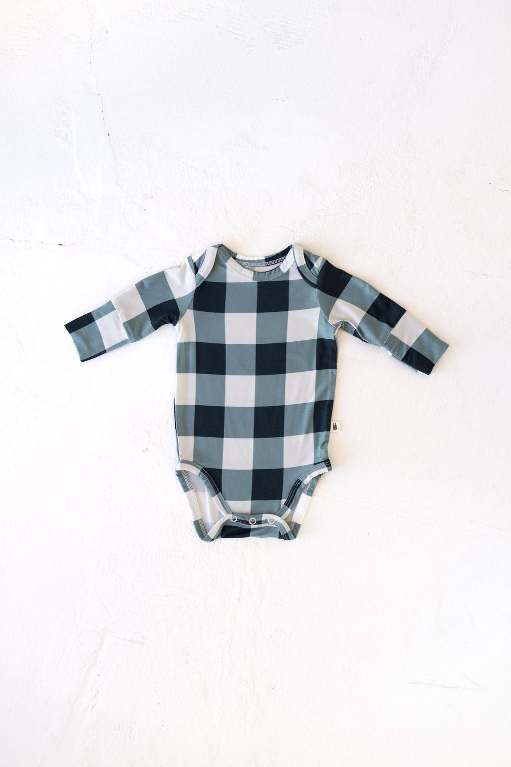 The "Long Sleeve Bamboo Snapsuit | Evergreen Gingham" by forever french baby is arranged flat on a light background. This black and white plaid baby onesie is crafted from breathable bamboo fabric, featuring a checkered pattern with hypo-allergenic qualities and snap closures at the bottom.