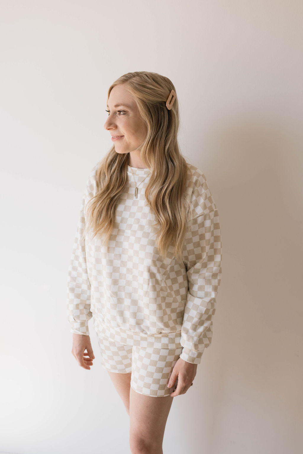 A person with long, wavy blonde hair stands in front of a plain white background, wearing the Adult Long Sleeve Short Set in Chai Checkerboard from forever french baby. They have a subtle smile and adorn their hair with a light-colored clip on the left side.