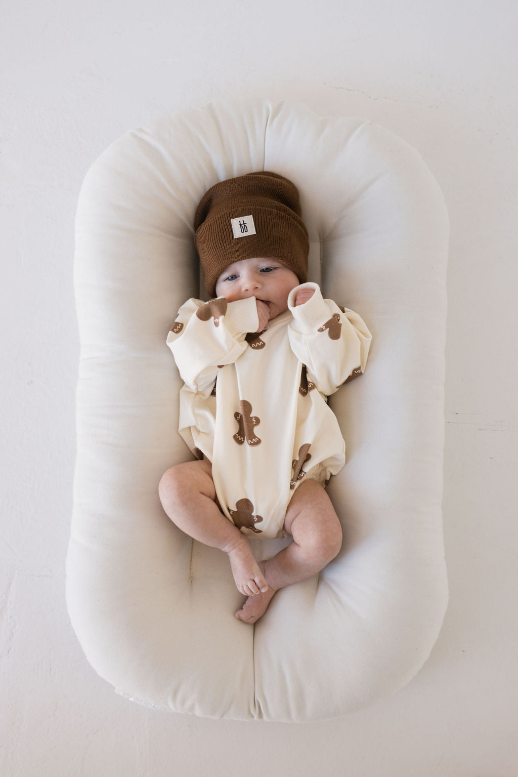 A baby wearing a Gingerbread Sweatshirt Romper from forever french baby lies on a white, cushioned surface. Their hands are near their face, and they seem cozy and relaxed in this adorable romper, perfect for the holiday season.