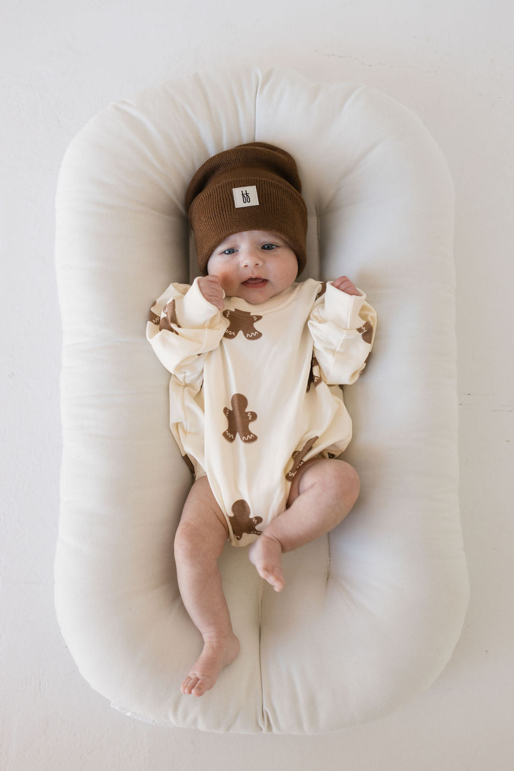 A baby lies on a cushioned surface, dressed in the forever french baby "Gingerbread" sweatshirt romper and wearing a brown knit hat. Perfect for the holiday season, this scene captures the warmth of winter and sweet moments of matching sets with loved ones.