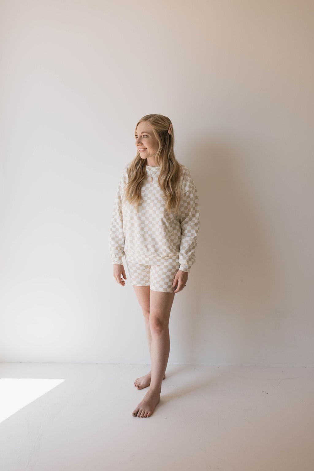 A blonde woman stands barefoot against a plain white background, wearing the "Adult Long Sleeve Short Set | Chai Checkerboard" by forever french baby. She gazes to her left with a slight smile, embodying the charm of matching long-sleeve short sets, arms relaxed by her side.