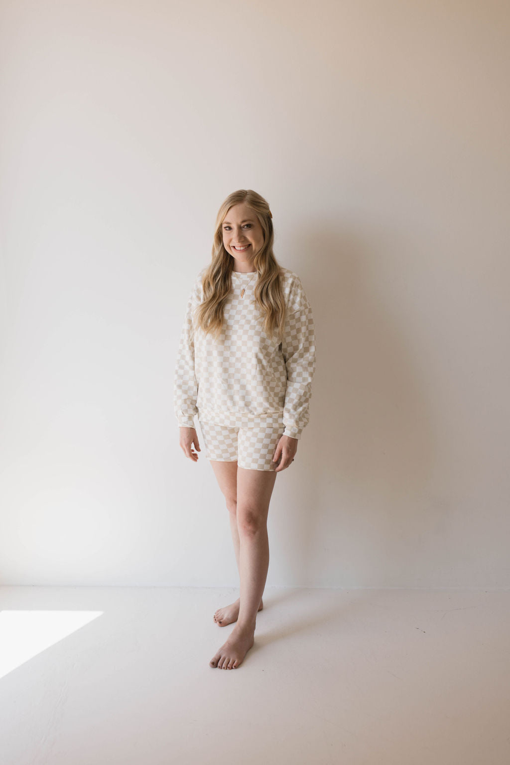 A person with long, blonde hair is standing barefoot against a neutral-colored wall. They are wearing the Adult Long Sleeve Short Set in Chai Checkerboard by forever french baby. Smiling warmly with their hands by their sides, they give off the perfect mama-and-me moment vibe in the softly lit room.