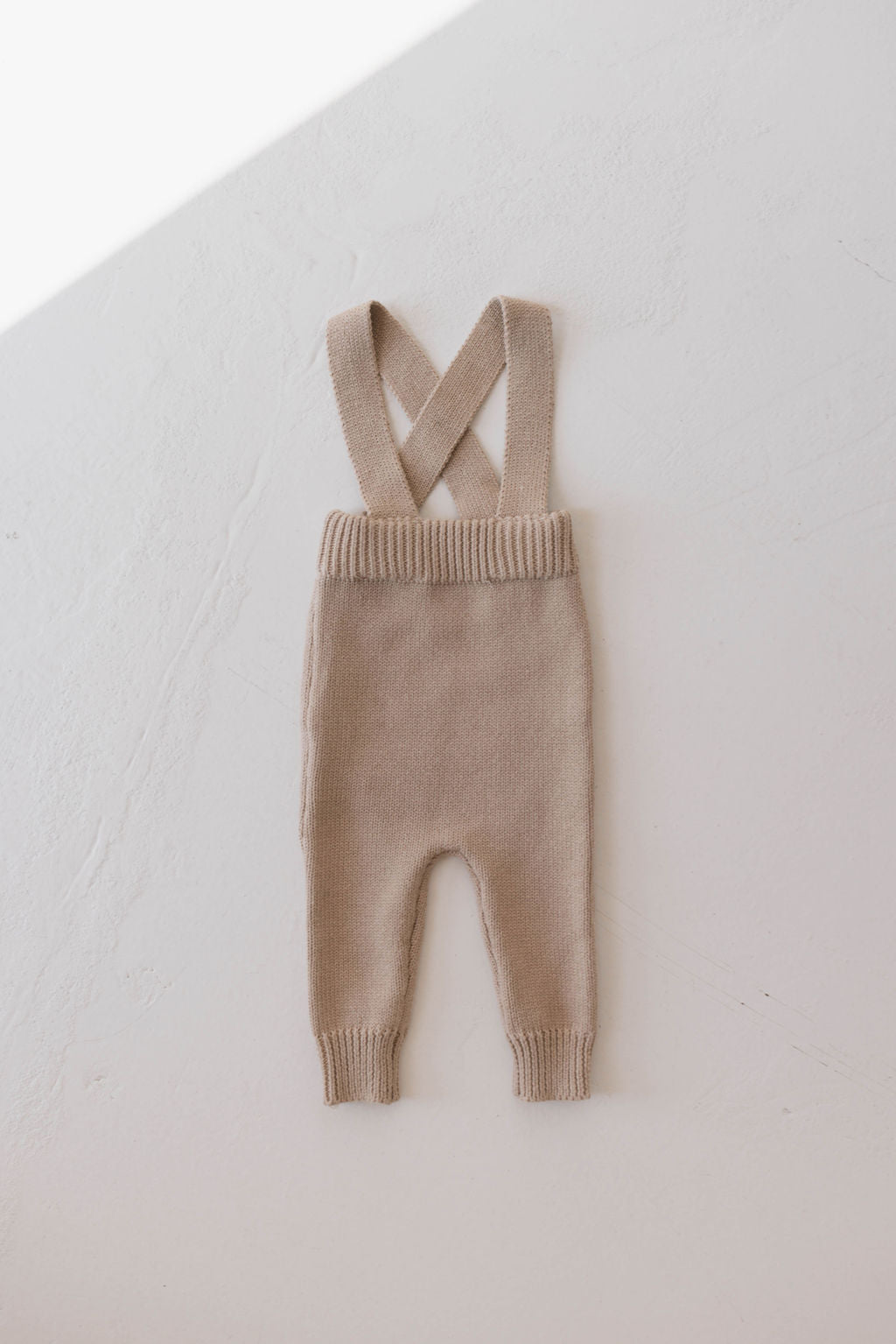 A pair of Graham Knit Suspenders by Forever French is laid flat against a white background. Perfect for cooler weather, these light brown baby overalls feature ribbed cuffs at the ankles and waist and have crossed straps at the top to form suspenders.