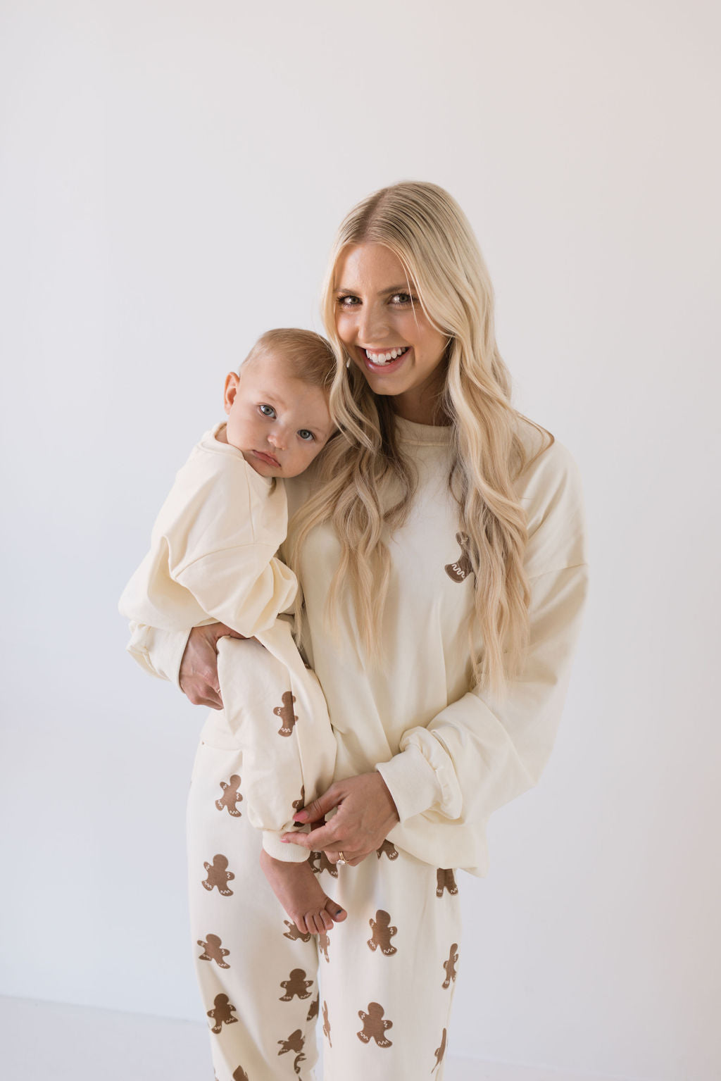 A woman with long blonde hair smiles while holding a baby, both dressed in matching family outfits from Forever French Baby's "Sweat Set | Gingerbread." Their cream-colored ensembles adorned with brown teddy bear patterns are ideal for the holiday season. The plain light backdrop accentuates their delightful bond.