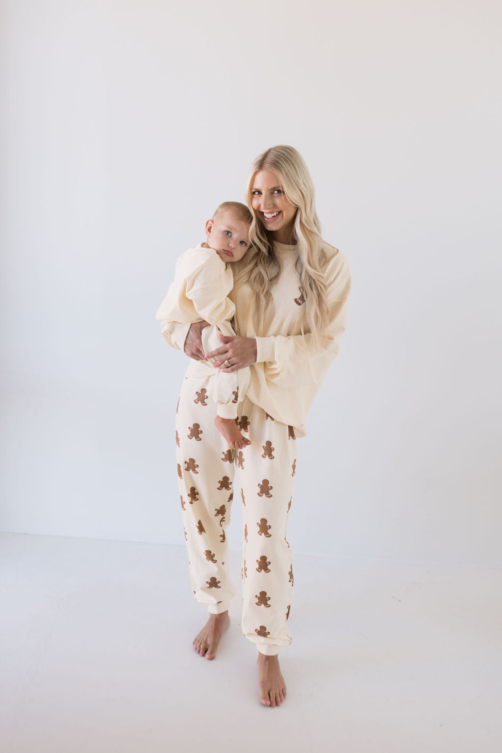 A woman with long blonde hair cradles a baby, both adorned in matching cream-colored outfits featuring gingerbread man patterns from the forever french baby Adult Sweat Set collection. This festive set perfectly captures the holiday spirit against a plain white backdrop.