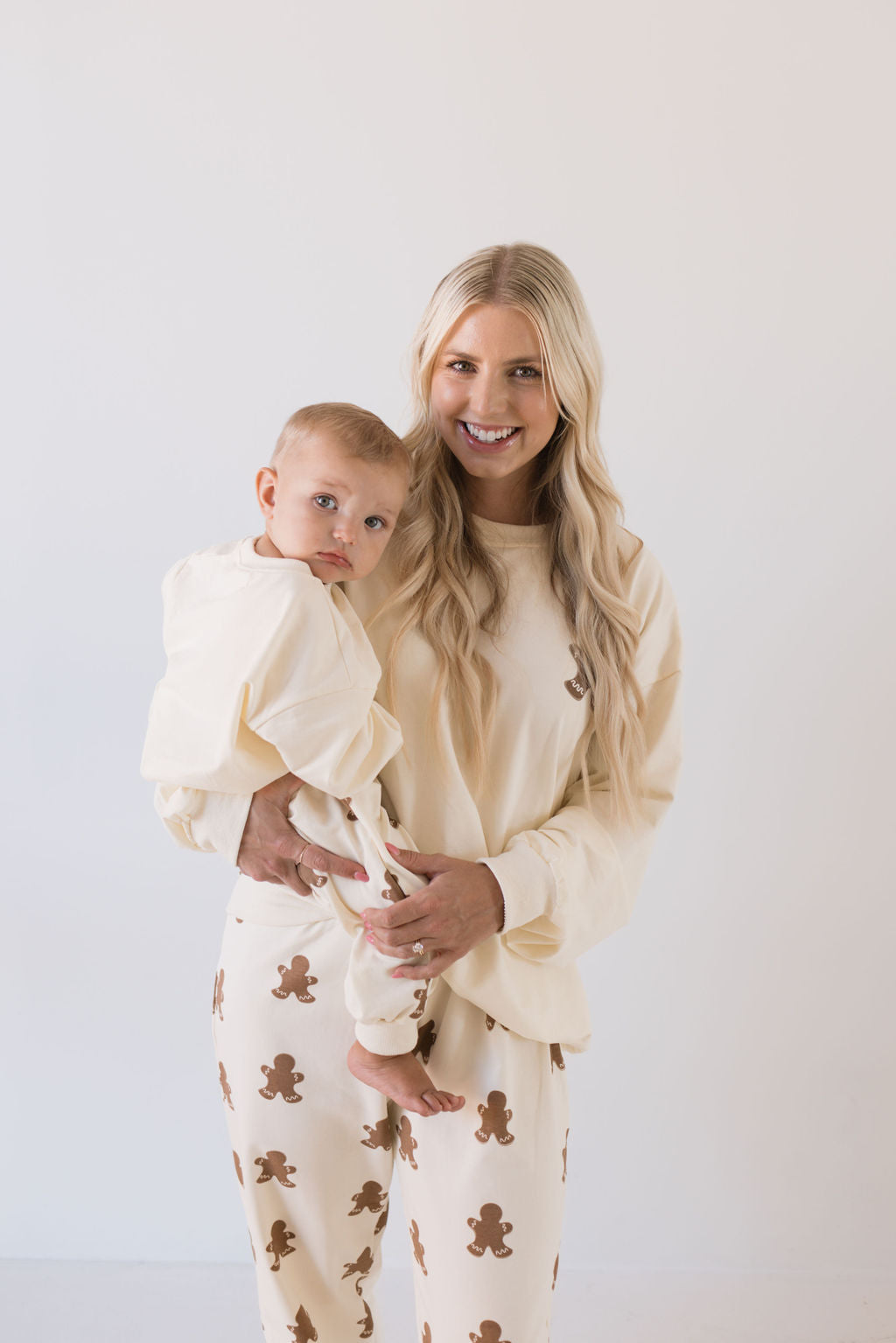 A woman with long blonde hair smiles while holding a baby. They are both dressed in matching outfits—hers is from the forever french baby collection, featuring the Adult Sweat Set in Gingerbread—capturing a cozy holiday spirit against a white background.