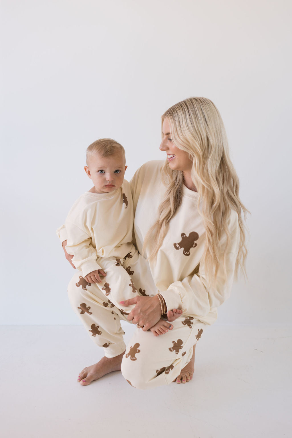 A woman with long blonde hair kneels and smiles at a toddler she is holding, both dressed in matching "Sweat Set | Gingerbread" outfits from forever french baby, featuring cream fabric adorned with charming brown teddy bear prints—ideal for the holiday season. They are set against a plain white background.