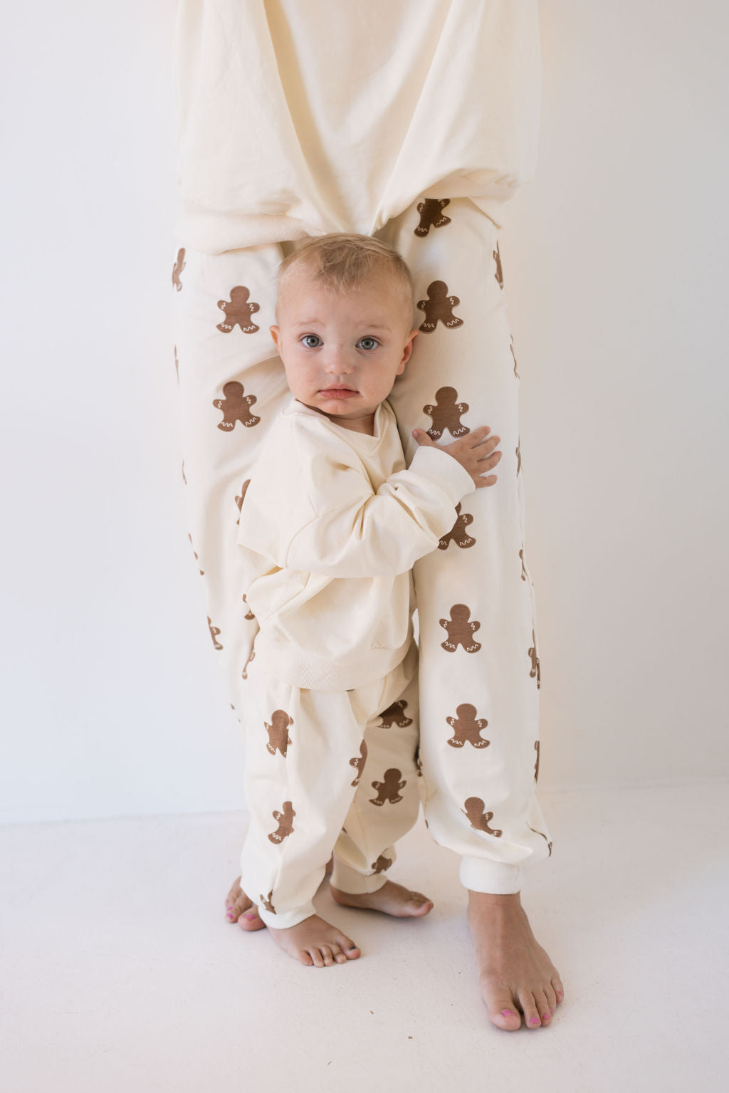 A toddler in cream-colored pajamas with brown bear patterns leans against an adult's legs, both dressed in the "Sweat Set | Gingerbread" from forever french baby. They're sporting matching family outfits, ideal for the holiday season. The plain, light background enhances the cozy and festive atmosphere.