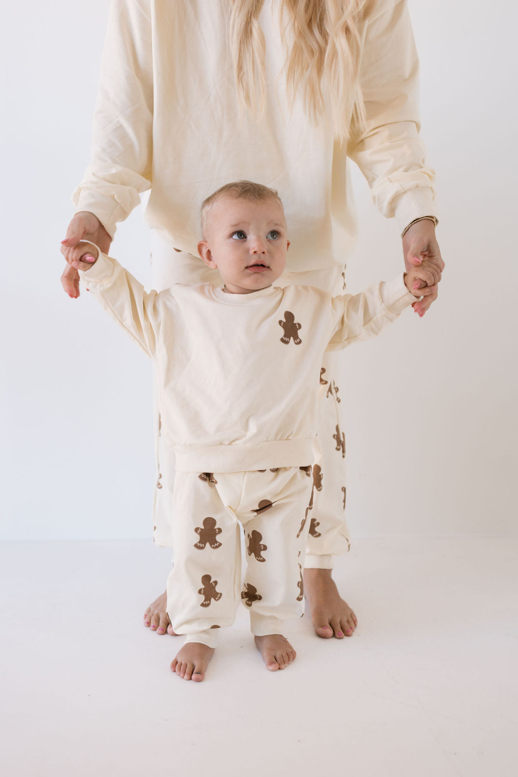 A toddler wearing creamy gingerbread-print pajamas from forever french baby stands while holding an adult's hands. Both are dressed in matching family outfits from the Sweat Set | Gingerbread collection, ideal for the holiday season, set against a plain white background with the adult's head not visible.