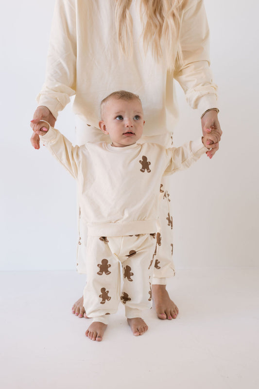 A toddler wearing creamy gingerbread-print pajamas from forever french baby stands while holding an adult's hands. Both are dressed in matching family outfits from the Sweat Set | Gingerbread collection, ideal for the holiday season, set against a plain white background with the adult's head not visible.