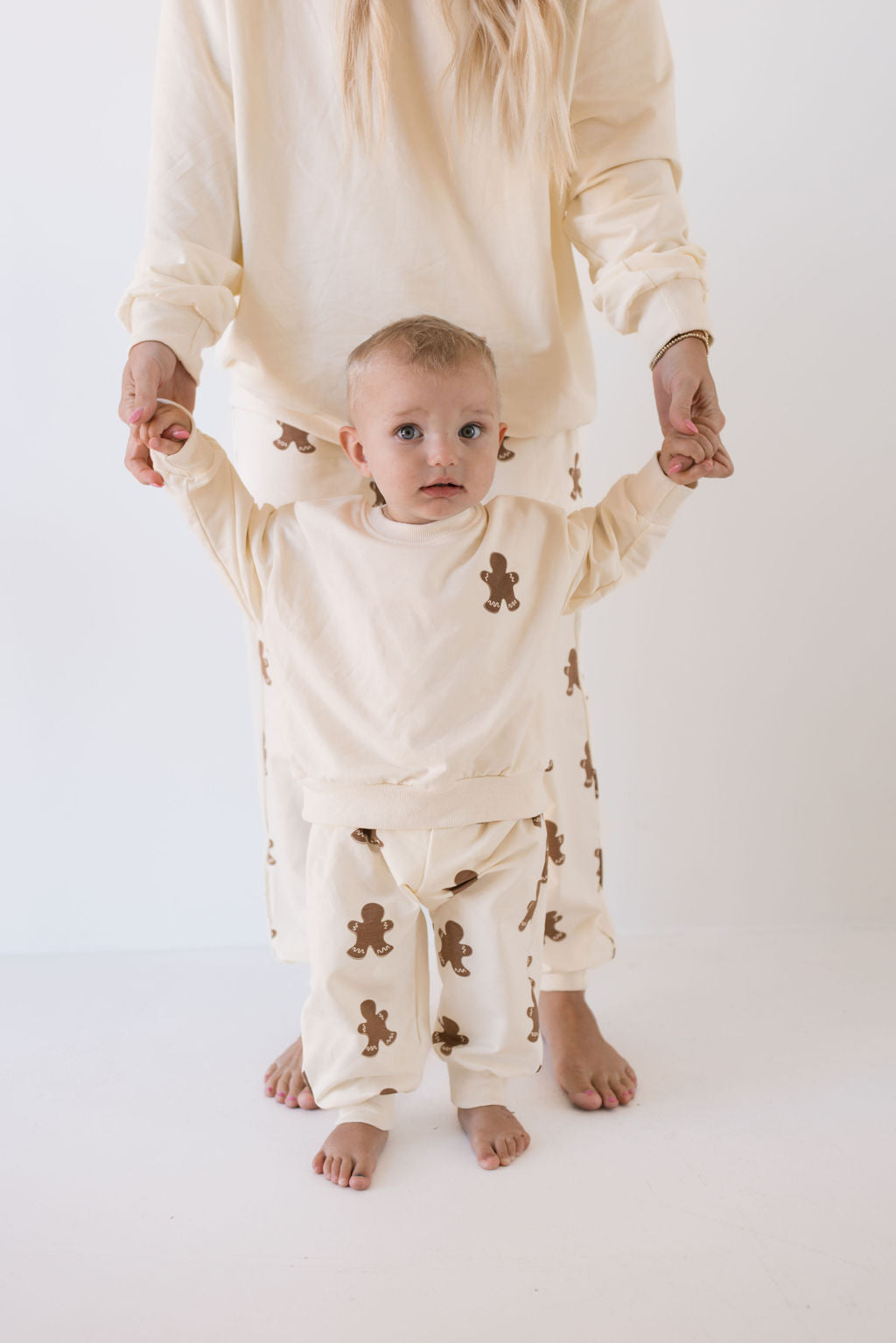 A baby and an adult, both dressed in matching 'Sweat Set | Gingerbread' outfits by forever french baby—cream-colored with brown teddy bear patterns—stand barefoot against a plain white background. Perfect for the holiday season, the adult holds the baby's hands for support.