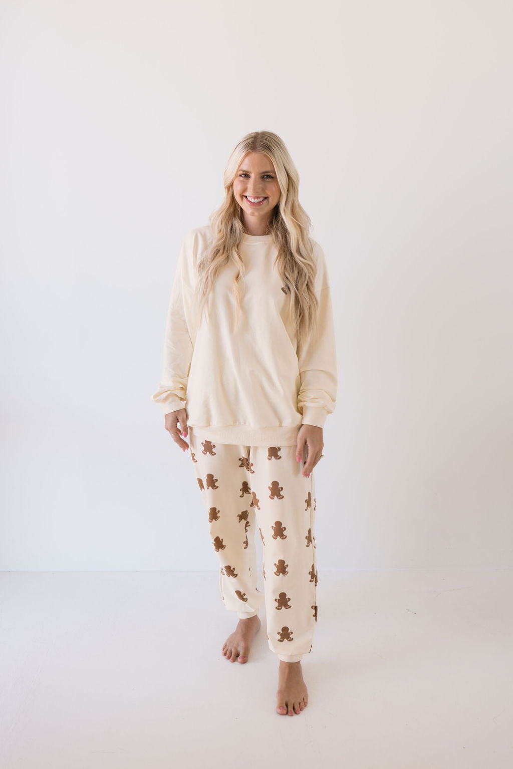 A woman with long blond hair, dressed in the cozy Adult Sweat Set | Gingerbread from forever french baby, smiles while standing barefoot on a white background. Perfect for the holiday season, her outfit exudes warmth and festive charm.