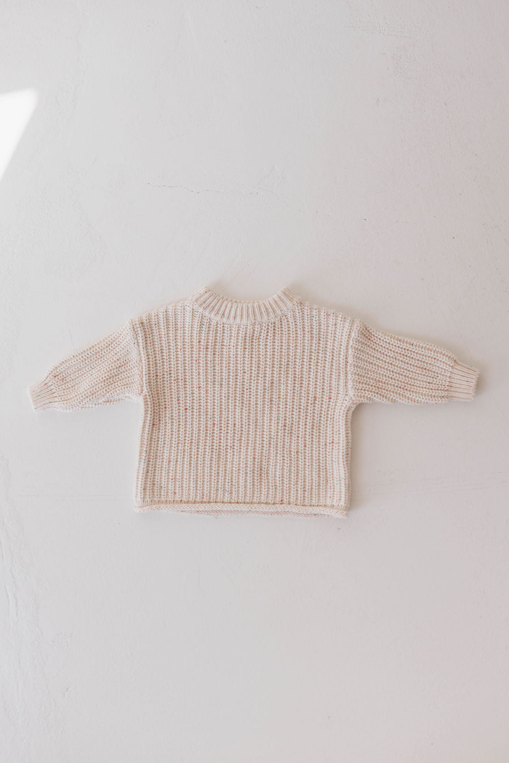 A small, beige knit sweater with long sleeves is displayed flat on a white, textured surface. This cozy baby clothing piece, from the Forever French Baby brand’s Funfetti collection, appears to be handmade and exudes a simple yet inviting design.