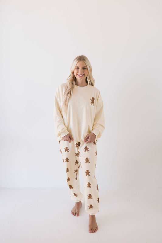 A person with long blonde hair is smiling in the *Adult Sweat Set | Gingerbread* by forever french baby, featuring a cream color with charming brown teddy bear patterns. Ideal for the holiday season, this cozy ensemble stands out beautifully against the plain white background.