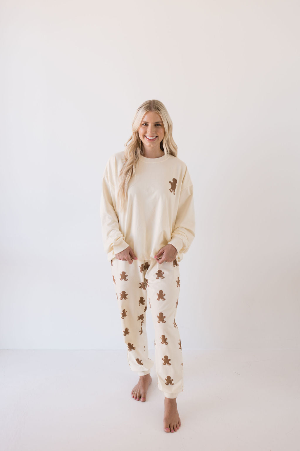 A woman beams in her Gingerbread Adult Sweat Set from forever french baby, perfect for the holiday season. She stands barefoot in a minimalist, white setting, embodying cozy elegance.