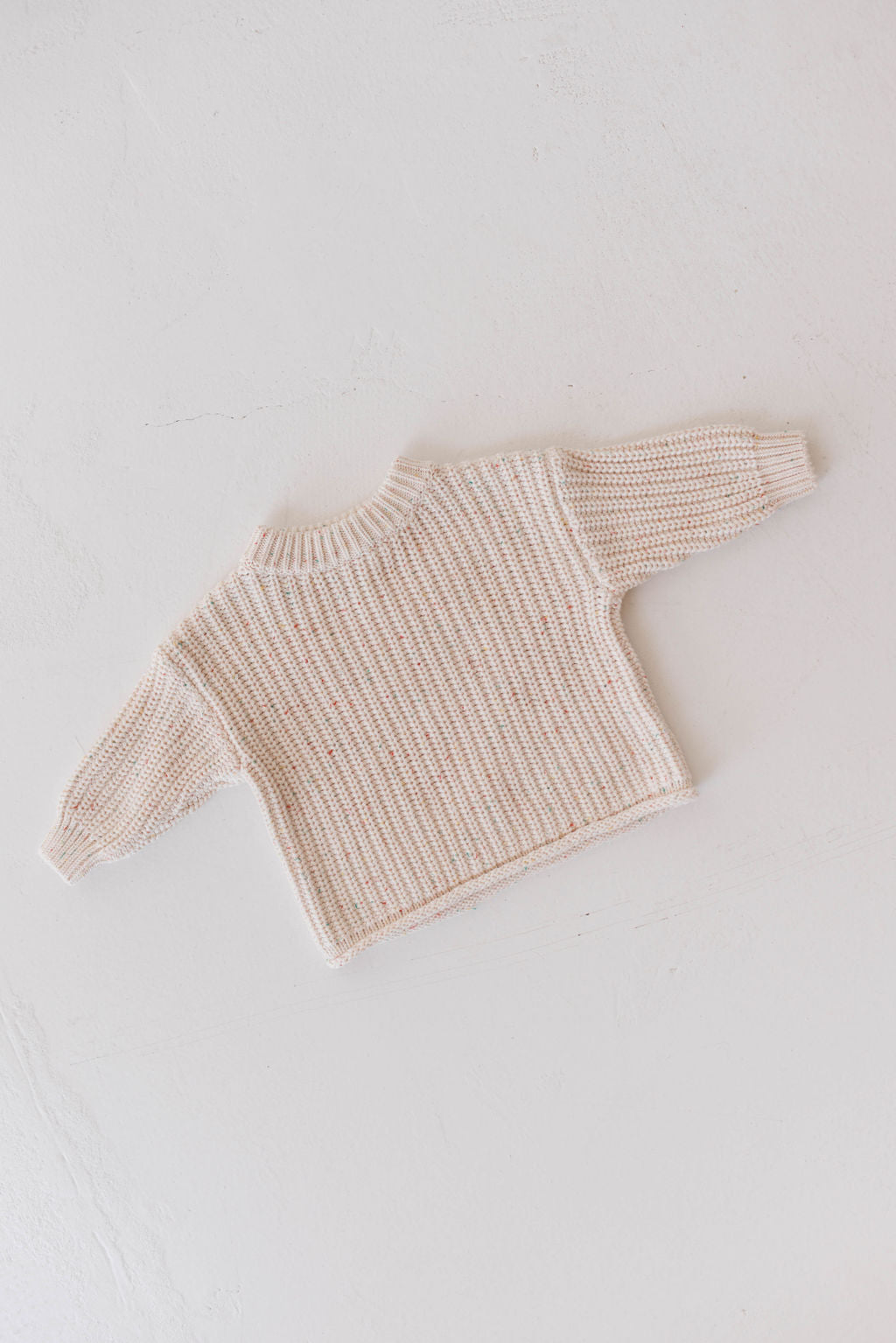 A small, cream-colored "Knit Sweater | Funfetti" by forever french baby lies flat on a light gray surface. The cozy baby sweater features a ribbed texture with long sleeves and a round neckline, appearing to be made from soft, comfortable material perfect for little ones.