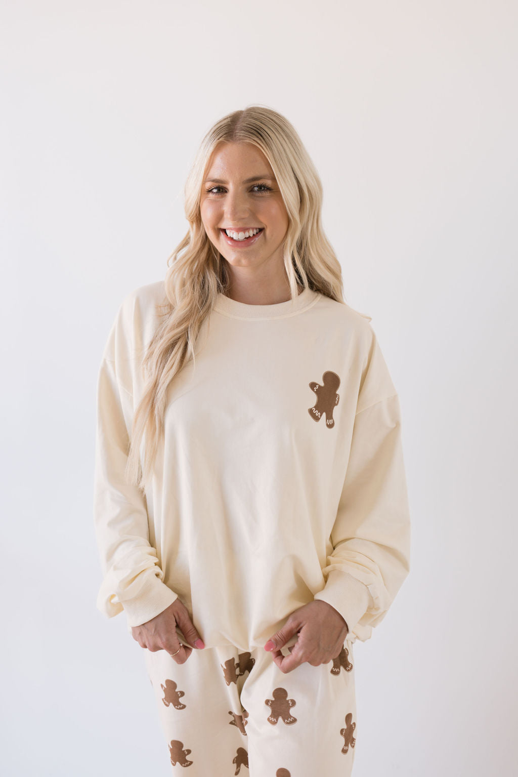 A person with long blonde hair smiles, dressed for the holiday season in an oversized cream-colored "Adult Sweat Set | Gingerbread" by forever french baby. The background is plain white.