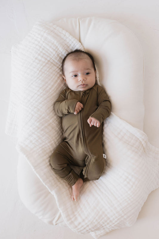 A baby dressed in olive green Bamboo Zip Pajamas from forever french baby is lying on a padded white blanket. The baby is looking up, with one hand slightly curled and the other resting on their chest. The minimal surrounding area enhances the focus on the baby in their premium bamboo pajamas from ff club.